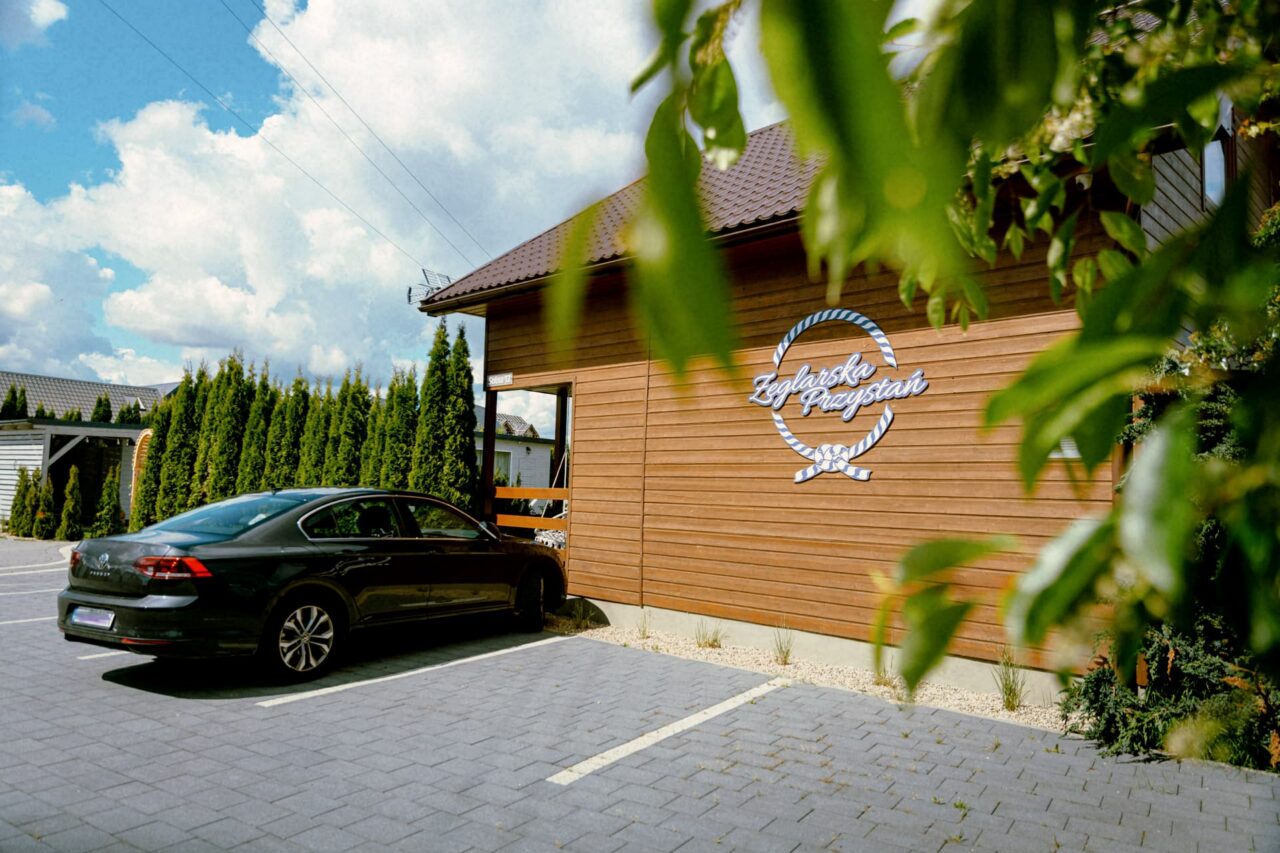 Samochód stoi na parkingu przy drewnianym budynku, którego ścianę oznacza duże logo Żeglarska Przystań.