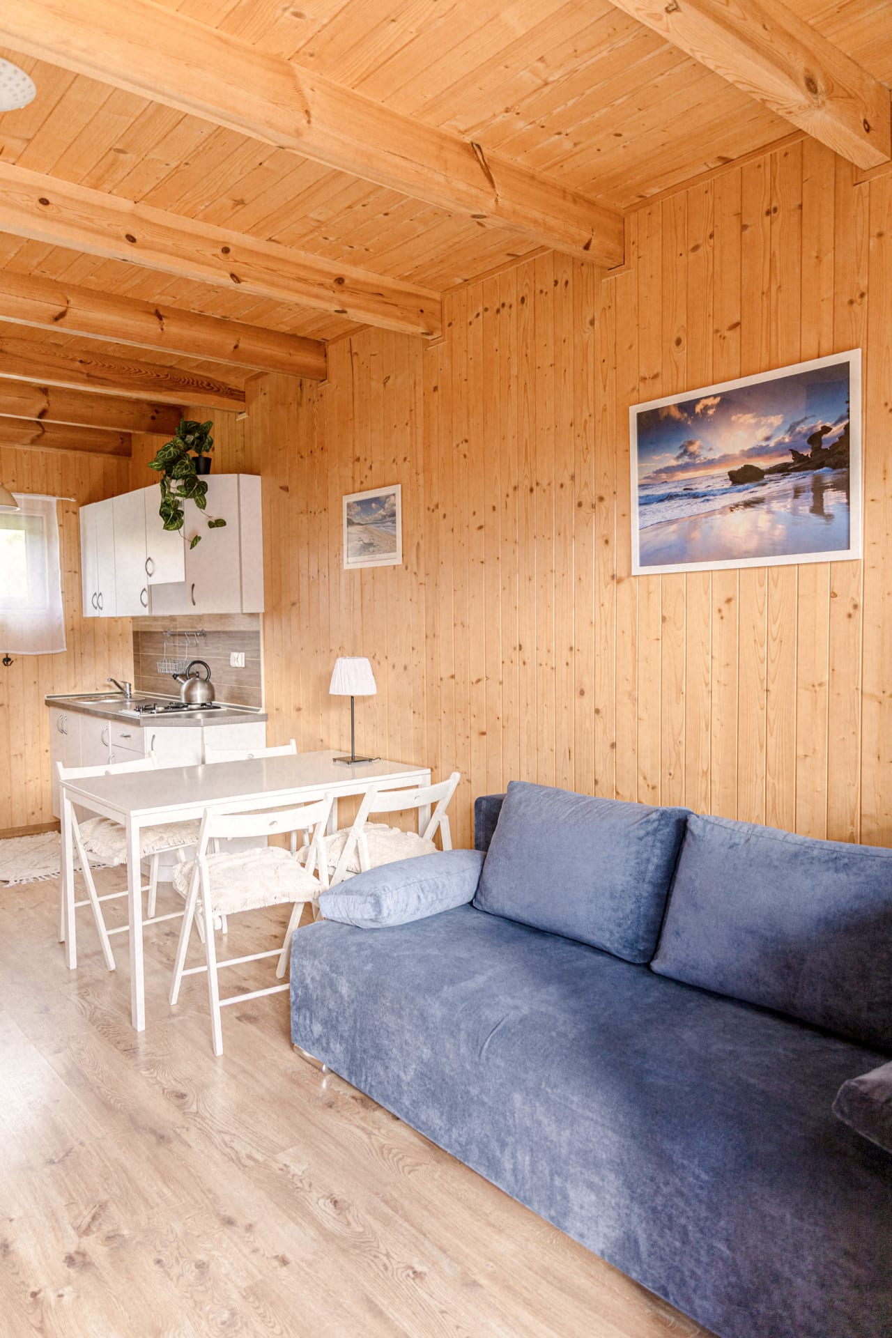 Niebieska sofa, biały stół i aneks kuchenny w salonie wykończonym naturalnym drewnem w domku letniskowym.