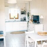 Jadalnia, telewizor i biały aneks kuchenny tworzą jasny salon w domku letniskowym w Chłapowie.
