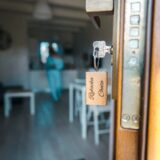Klucz z drewnianym brelokiem i napisem „Rybacka Chata” w otwartych drzwiach i rozmytym wnętrzem domku letniskowego Żeglarska Przystań.